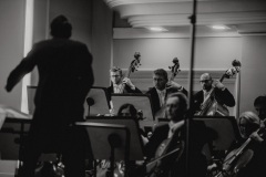 Czarno-białe zdjęcie. Sekcja wiolonczel i kontrabasów, dyrygent stojący tyłem.