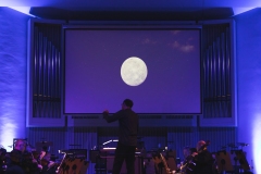 Widok na scenę podświetloną niebieskimi światłami, na ekranie grafika przedstawiająca planetę